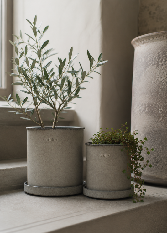 Two matte gray concrete plant pots with matching saucers, one large and one small, displayed on a stone windowsill next to a textured ceramic vase.