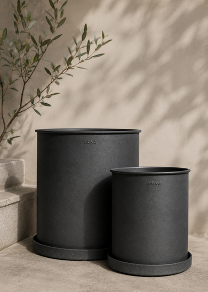 Two matte black cylindrical planters with matching saucers, one large and one small, displayed on a concrete surface with a warm beige wall and soft leaf shadows in the background.